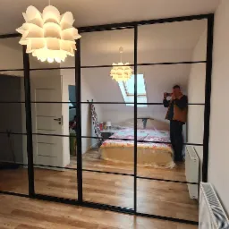 Czarna szafa wnękowa z lustrzanymi drzwiami przesuwnymi, podzielonymi na kwadratowe panele, odbijająca widok pokoju z ł&oacute;żkiem na poddaszu i osobę robiącą zdjęcie telefonem.