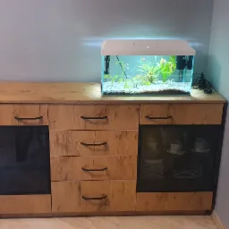 Komoda z frontami imitującymi drewno, z czarnymi uchwytami i przeszklonymi witrynami, na której stoi akwarium z roślinami i rybkami, obok żółte zasłony.