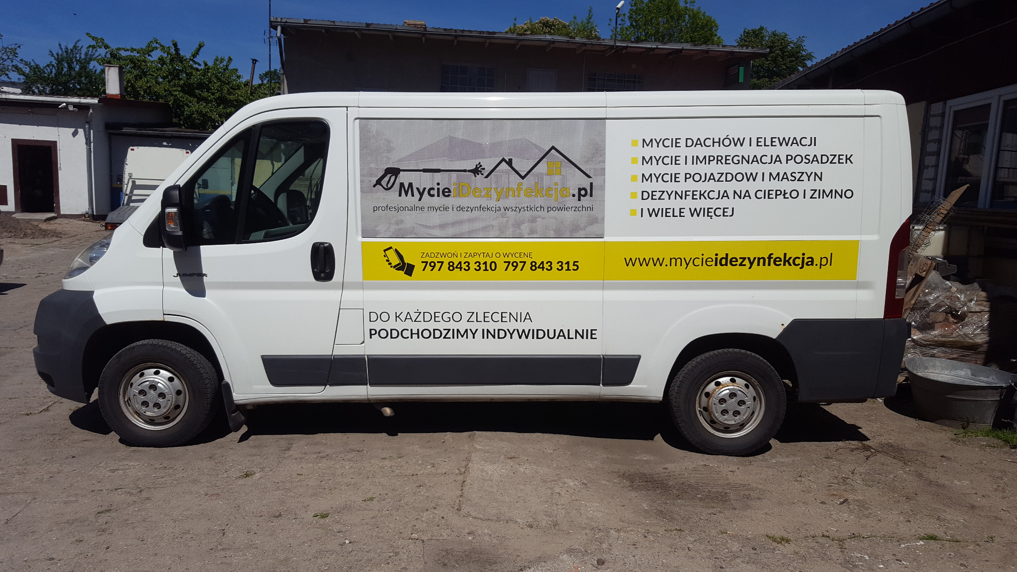 Biały samochód dostawczy z reklamą firmy myjącej dachy, elewacje, posadzki oraz oferującej dezynfekcję, zaparkowany na zewnątrz budynku.