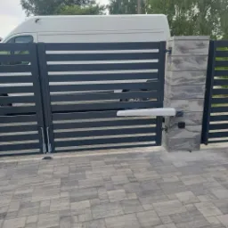 Nowoczesna, pozioma brama wjazdowa z automatyką, szara, z betonowymi słupkami, na tle białego samochodu dostawczego i kostki brukowej.