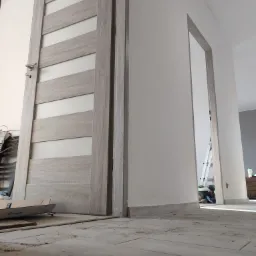 Montaż szarej, drewnopodobnej ościeżnicy z przeszklonymi panelami w trakcie prac wykończeniowych, widoczny karton i narzędzia na podłodze z szarych płytek, w tle drabina oparta o ścianę.