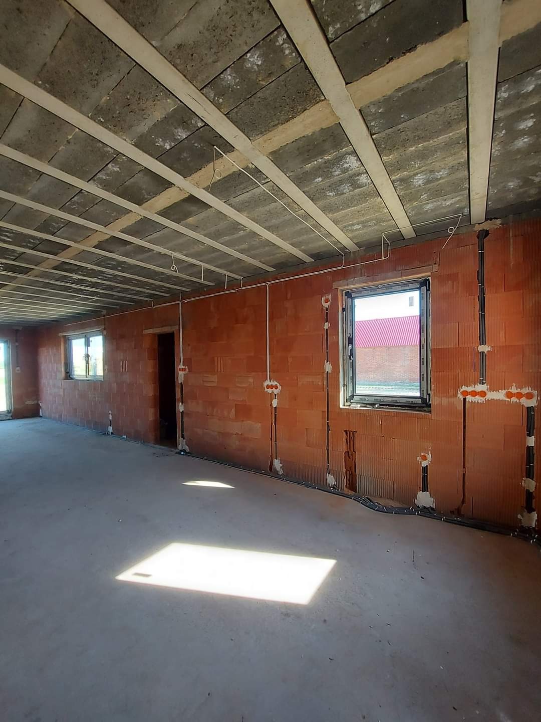 Surowy stan wnętrza budynku z widoczną instalacją elektryczną w bruzdach ceglanej ściany, betonowym stropem z drewnianymi belkami i oknami z widokiem na czerwony dach.