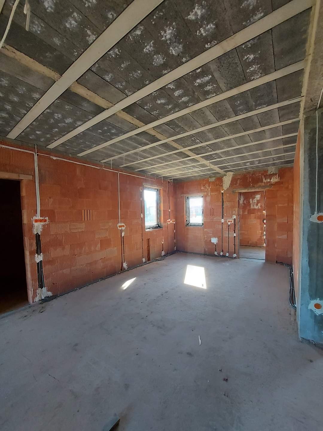 Surowy stan wnętrza: ściany z czerwonej cegły dziurawki, sufit betonowy z widocznymi belkami, instalacje elektryczne wyprowadzone ze ścian, posadzka betonowa w trakcie wylewania.