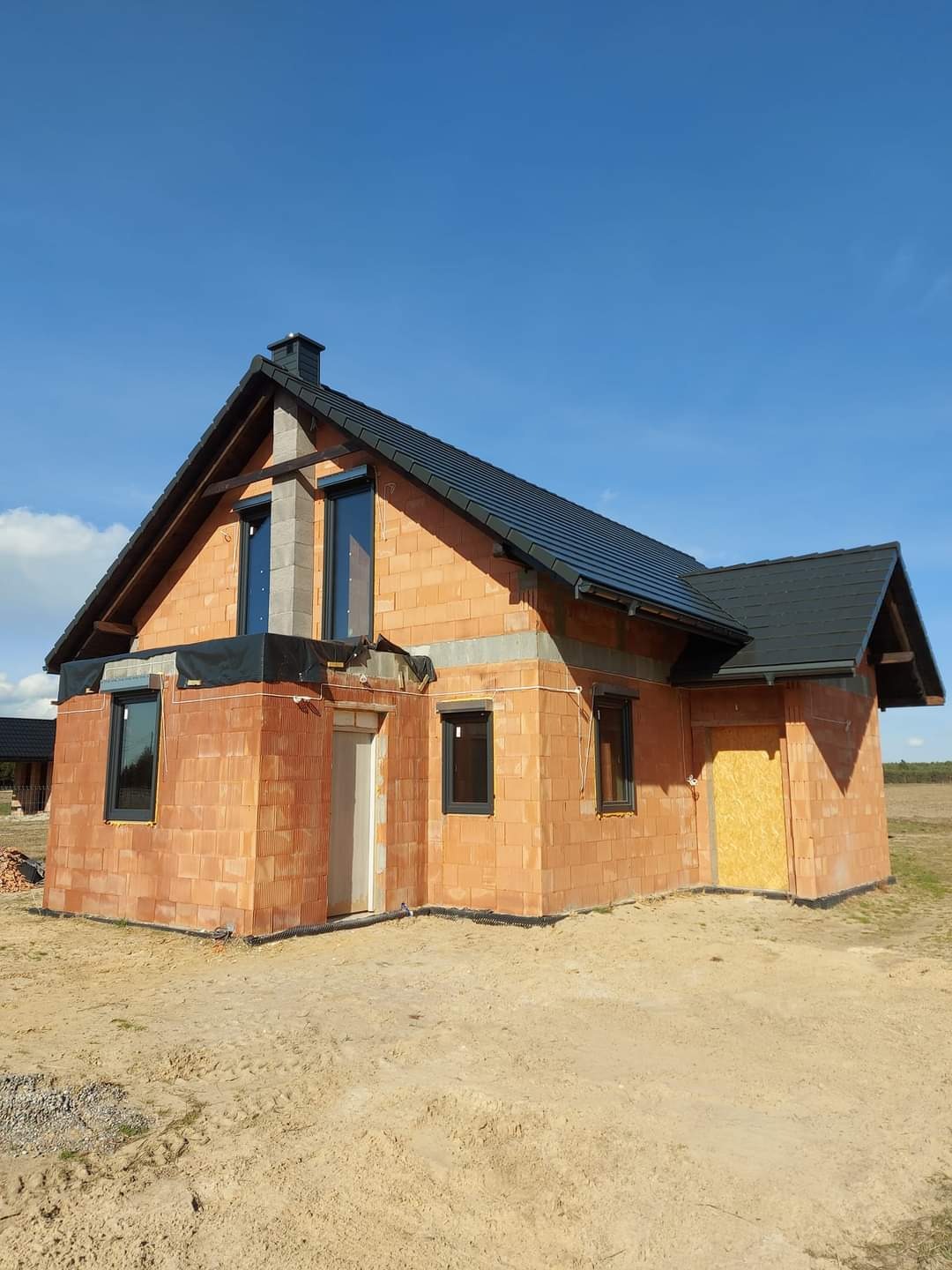 Surowy stan domu jednorodzinnego z czerwonej cegły z czarnym dachem i oknami, widoczne szare elementy konstrukcyjne, teren wokół domu piaszczysty.