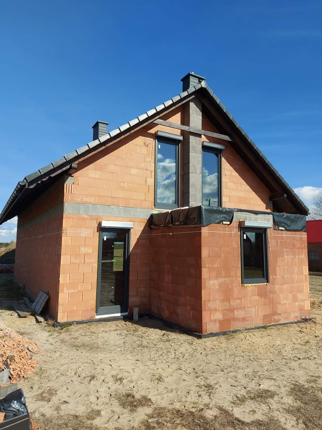 Surowy stan budynku jednorodzinnego z czerwonej cegły, z zamontowanymi oknami i roletami zewnętrznymi, widoczne elementy konstrukcyjne dachu i komina, teren wokół w stanie przygotowania...