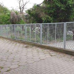 Wytwórnia i Wyroby z Drutu Łukasz Kraska - Szara siatka ogrodzeniowa z metalowymi słupkami na tle zieleni, widok z zewnątrz posesji, kostka brukowa na pierwszym planie.
