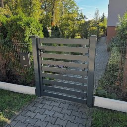 Wytwórnia i Wyroby z Drutu Łukasz Kraska - Nowoczesna, szara furtka z poziomych paneli, zamykana na czarny zamek, wkomponowana w siatkę ogrodzeniową. Obok skrzynka pocztowa i chodnik z kostki.