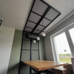 Wytwórnia i Wyroby z Drutu Łukasz Kraska - Nowoczesna, metalowa konstrukcja z siatki, zamontowana na suficie i ścianie, tworząca unikalny element dekoracyjny wnętrza z drewnianym stołem.