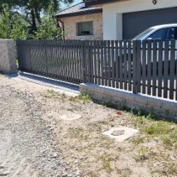 Nowoczesna, przesuwna brama wjazdowa w kolorze grafitowym z pionowymi sztachetami, zamontowana na posesji z kostki brukowej i elementami zieleni.