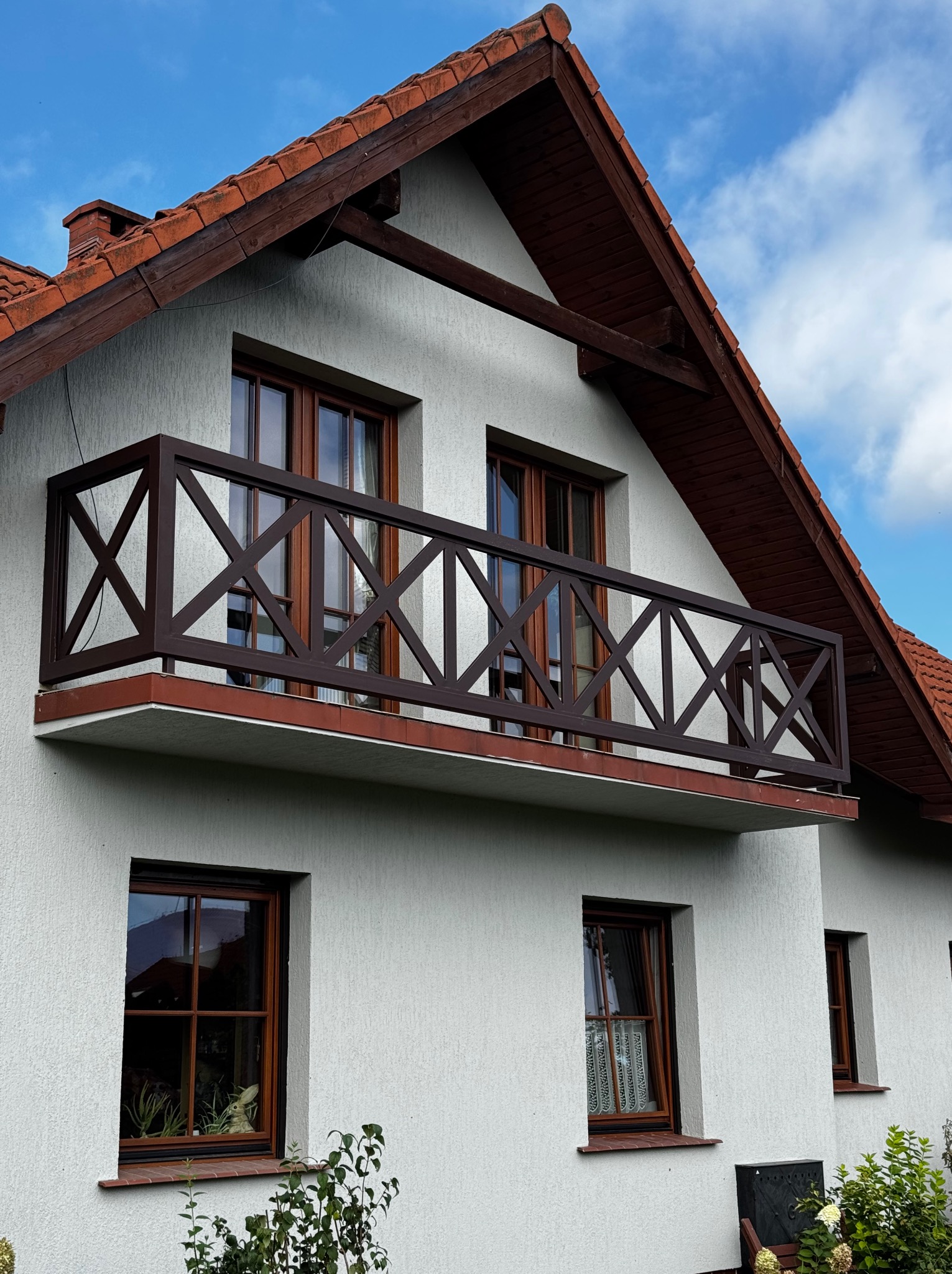 Elewacja domu z balkonem z brązową, drewnianą balustradą w kratkę. Dach z czerwonej dachówki. Biała, chropowata fasada budynku.