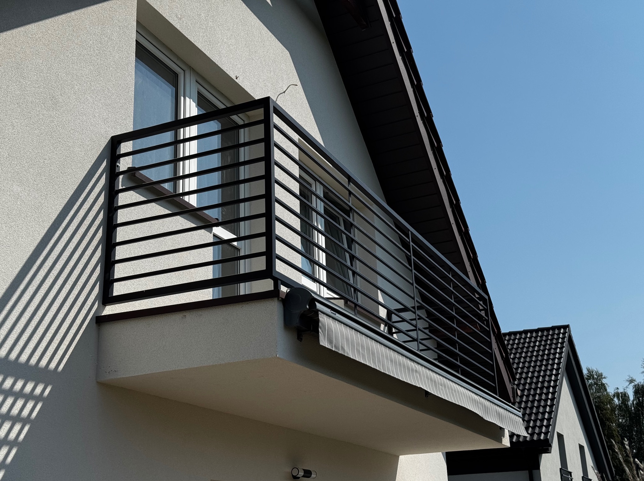 Nowoczesna, czarna balustrada balkonowa z poziomych profili, zamontowana na balkonie budynku z jasną elewacją. Widoczny fragment okna i zwinięta markiza.