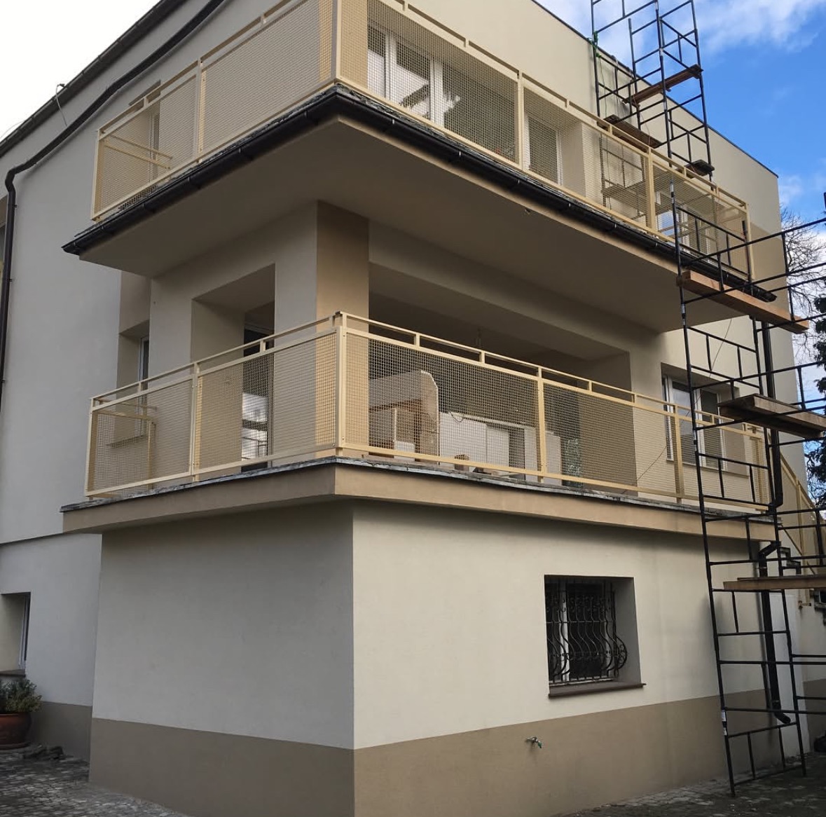 Elewacja budynku z beżowymi balustradami siatkowymi na balkonach, widoczne rusztowanie z boku. Ujęcie z dołu, błękitne niebo w tle.