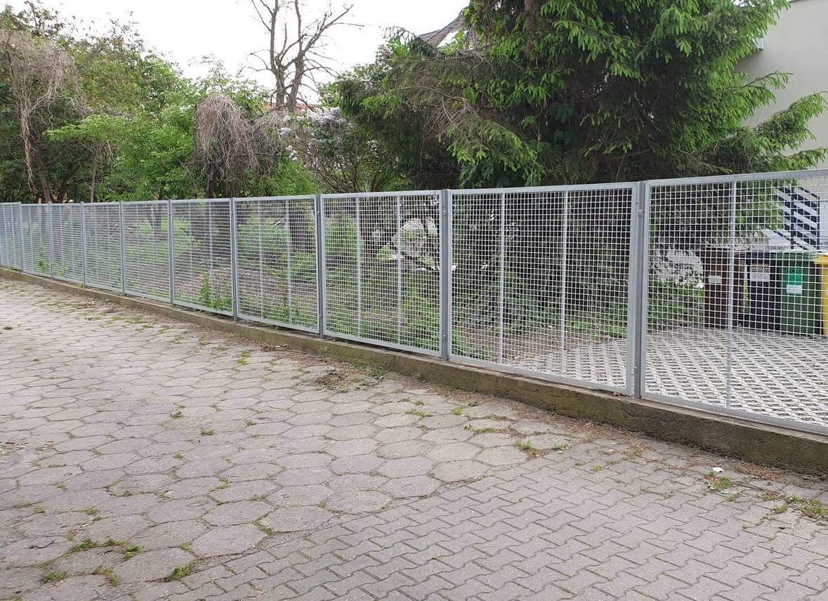 Szara siatka ogrodzeniowa z metalowymi słupkami na tle zieleni, widok z zewnątrz posesji, kostka brukowa na pierwszym planie.