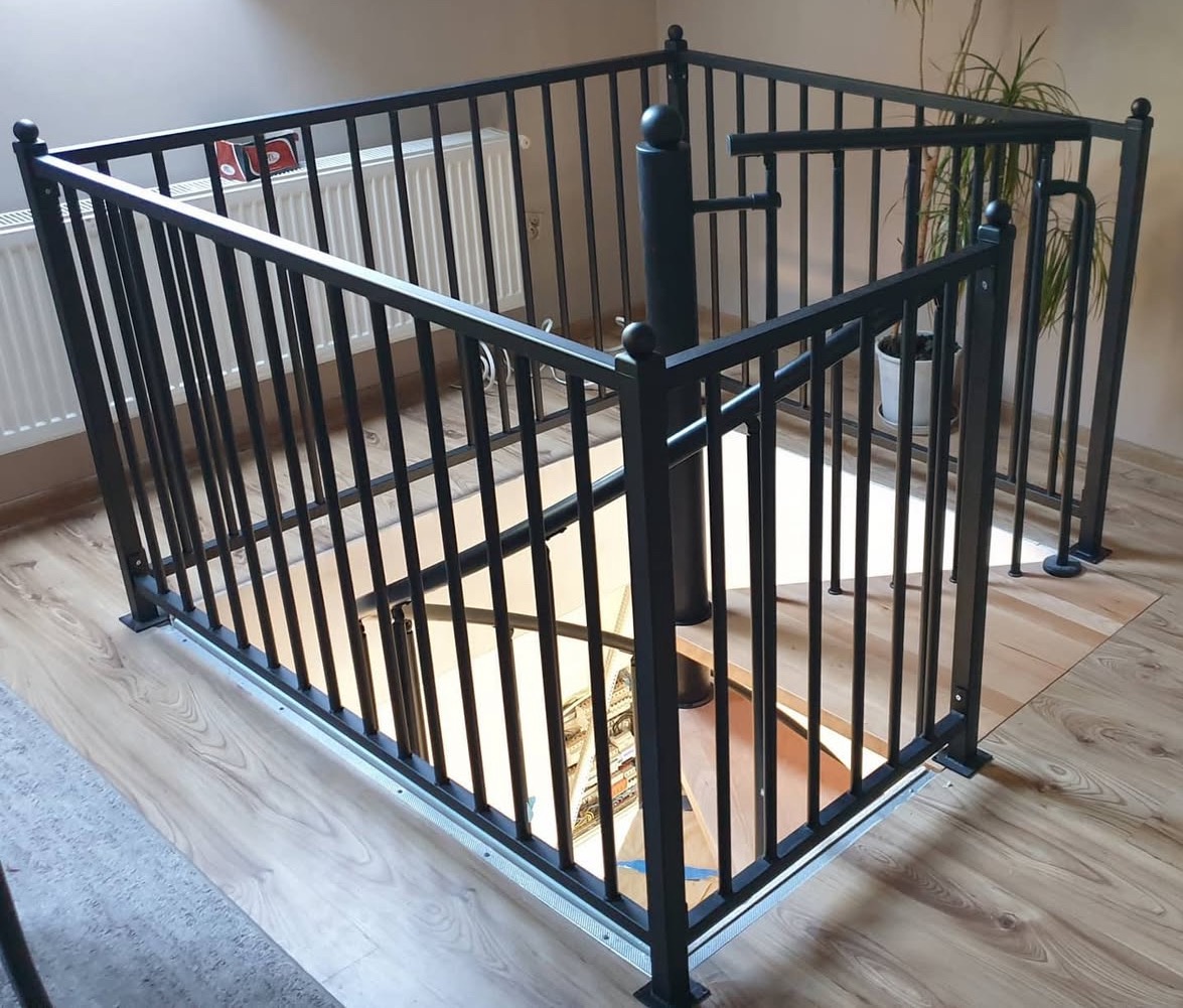 Kwadratowa, czarna balustrada zabezpieczająca otwór schodowy na drewnianej podłodze. Widoczny fragment schodów spiralnych i grzejnik.