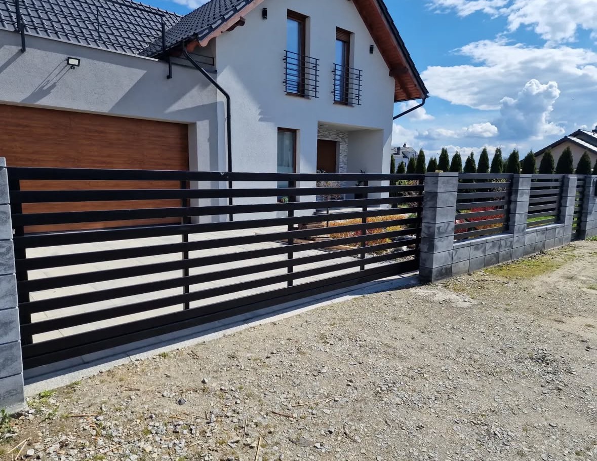 Nowoczesna, przesuwna brama wjazdowa z czarnymi, poziomymi profilami i szarymi, betonowymi słupkami na tle białego domu z brązową bramą garażową.