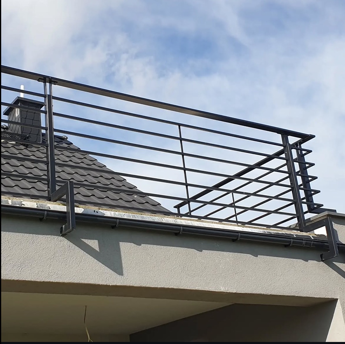 Nowoczesna, metalowa balustrada balkonowa w kolorze grafitowym na tle błękitnego nieba i dachu z dachówki. Minimalistyczny design z poziomymi prętami.