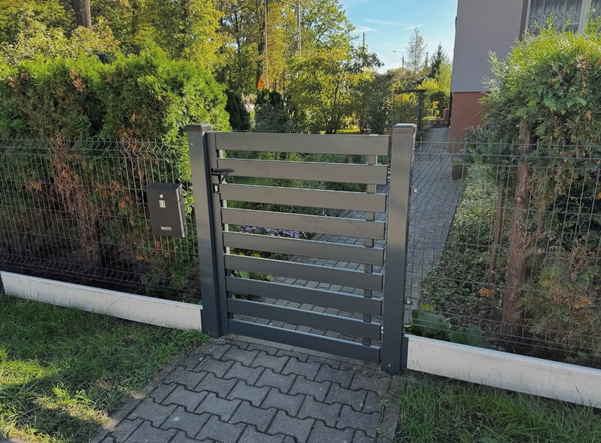 Nowoczesna, szara furtka z poziomych paneli, zamykana na czarny zamek, wkomponowana w siatkę ogrodzeniową. Obok skrzynka pocztowa i chodnik z kostki.