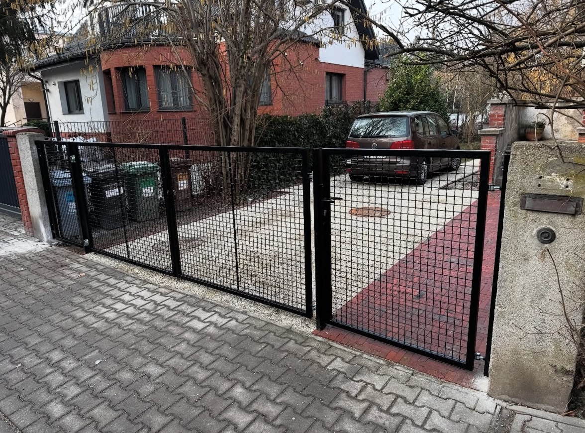 Czarna, metalowa brama z siatki kwadratowej, zamykająca wjazd na posesję z kostką brukową, z widocznym samochodem w tle i fragmentem domu z czerwonej cegły.