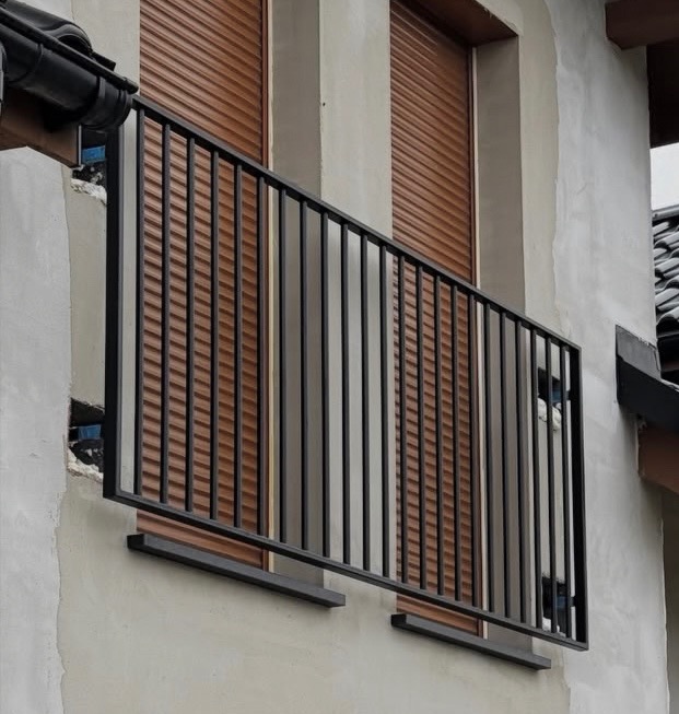 Czarna, metalowa balustrada balkonowa z pionowymi prętami, zamontowana na szarej elewacji budynku z brązowymi roletami w tle. Widok z dołu, lekko z boku.