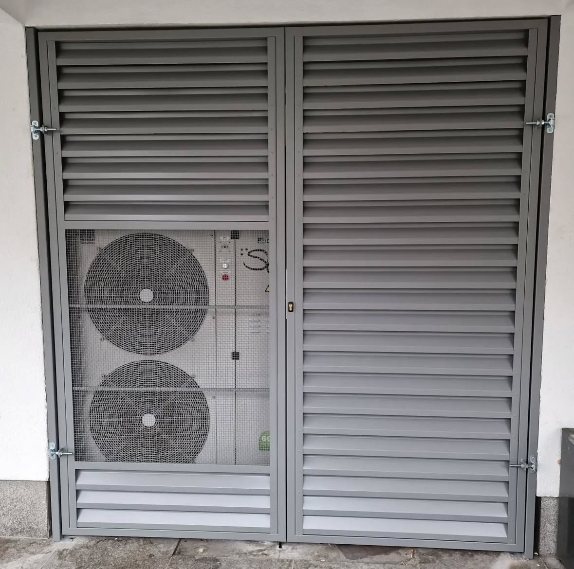 Szara, metalowa konstrukcja z żaluzjami i siatką maskującą agregat klimatyzacji na zewnątrz budynku. Widoczne zawiasy i zamek.