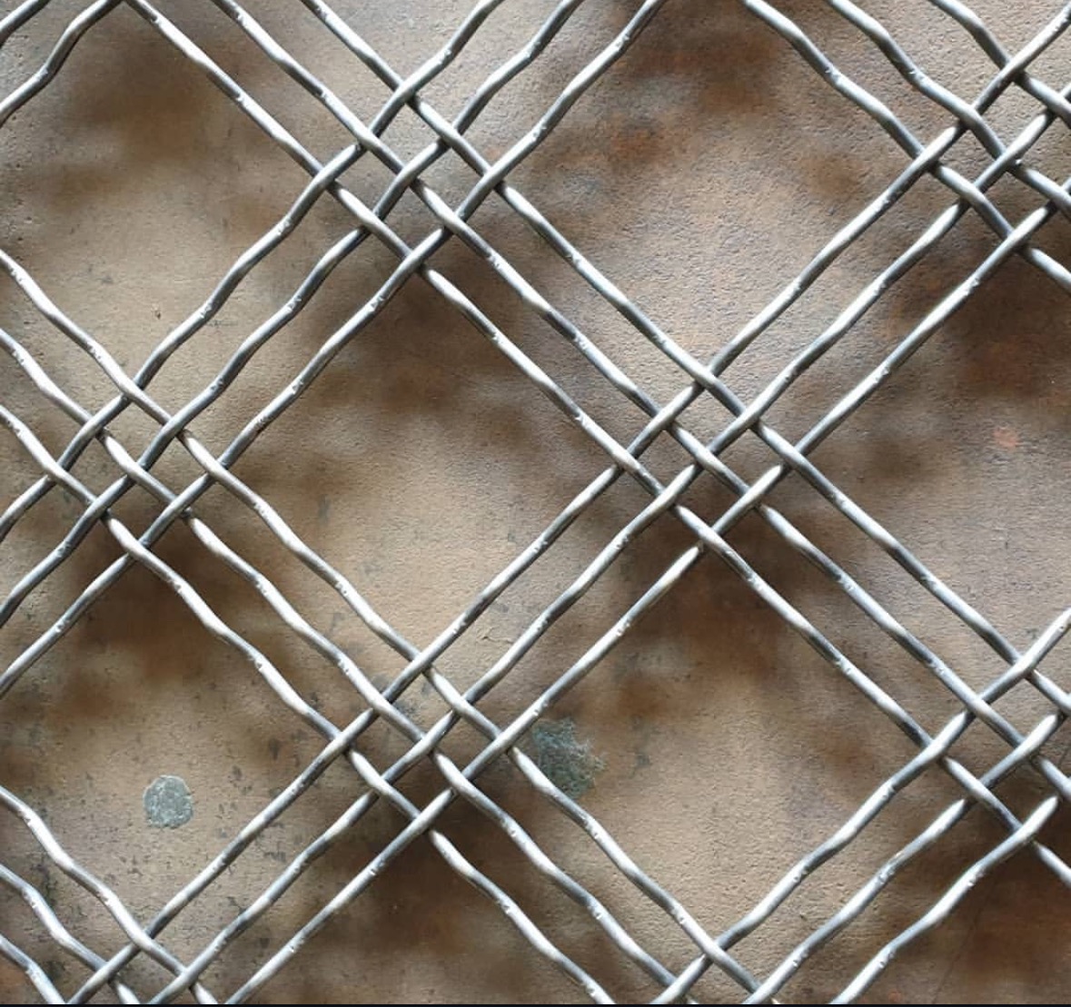 Metalowa siatka zgrzewana o romboidalnych oczkach na tle szarej ściany, zbliżenie na strukturę materiału.