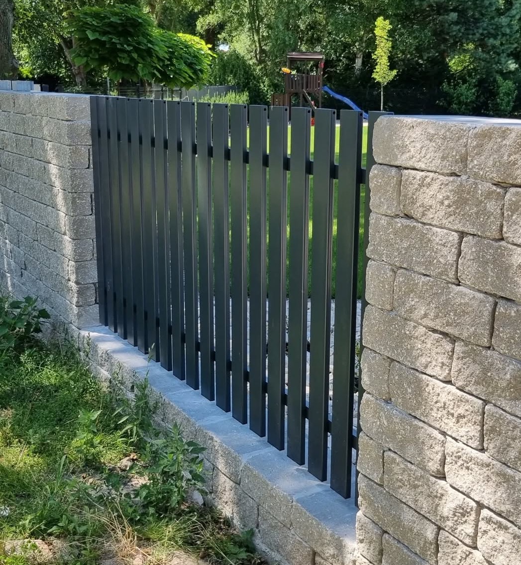 Nowoczesne, grafitowe ogrodzenie metalowe z pionowymi sztachetami, osadzone na murku z kamienia łupanego. Tło stanowi zielony trawnik i drzewa.