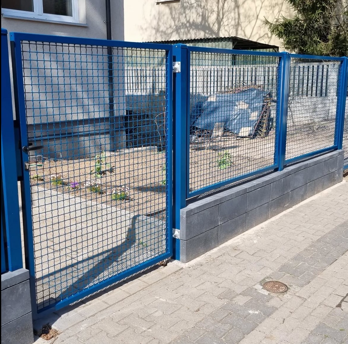 Niebieska, metalowa siatka ogrodzeniowa z bramką, osadzona na betonowej podmurówce. Widok z zewnątrz posesji na fragment chodnika i domu w tle.