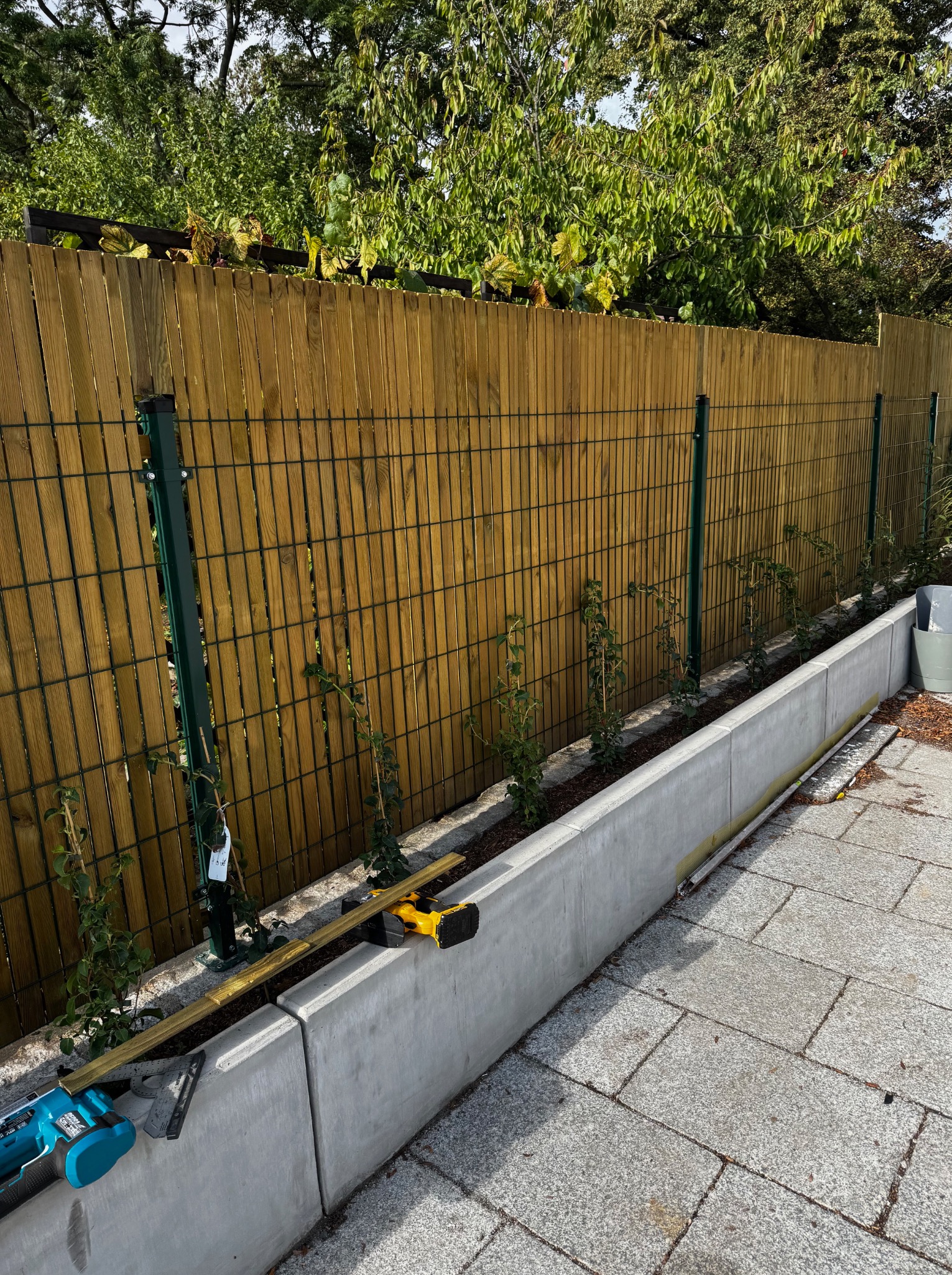 Panelowe ogrodzenie z drewnianym wypełnieniem, zielone słupki, betonowa podmurówka i narzędzia montażowe. Widok z boku na fragment ukończonego ogrodzenia.