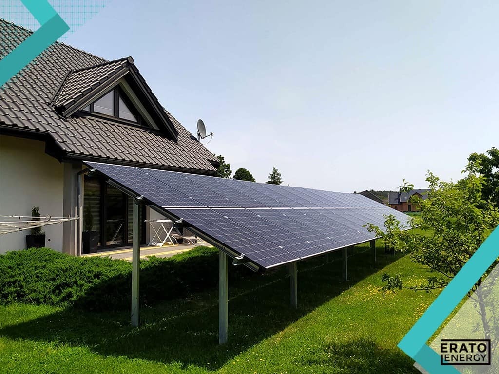 Instalacja paneli słonecznych na trawniku przed domem jednorodzinnym z ciemną dachówką, widoczne metalowe podpory i cień rzucany przez panele.