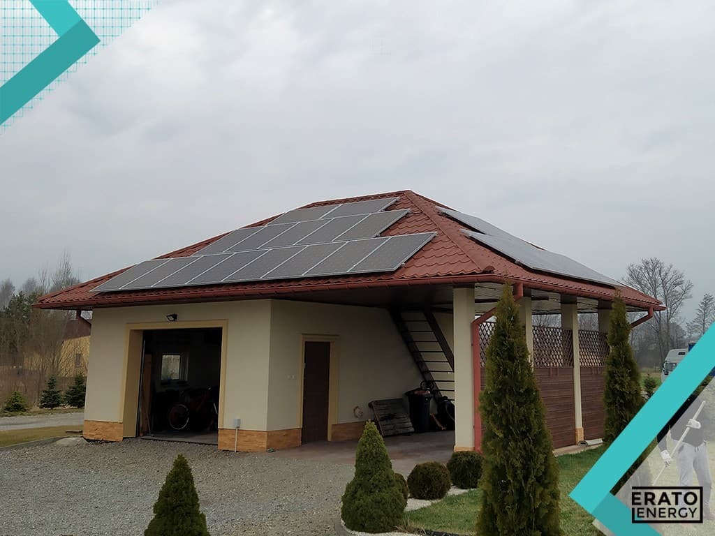Panele słoneczne na dachu budynku gospodarczego z otwartym garażem, widoczne tuje i logo firmy w rogu zdjęcia.
