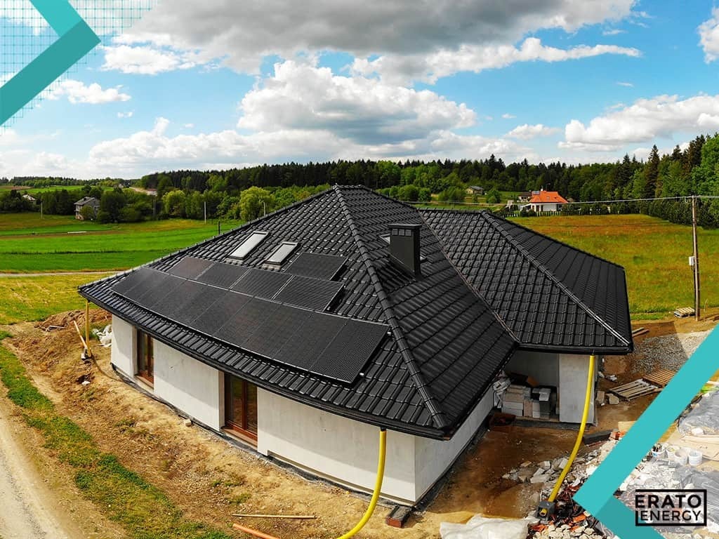 Dom jednorodzinny z zamontowanymi panelami słonecznymi na dachu pokrytym czarną dachówką, widok z góry, na tle zielonego krajobrazu i błękitnego nieba z chmurami.