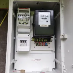 Otwarta skrzynka elektryczna z zamontowanym licznikiem energii, bezpiecznikami i elementami przyłączeniowymi. Na dnie skrzynki widoczne drobne narzędzia i plastikowy pojemnik.