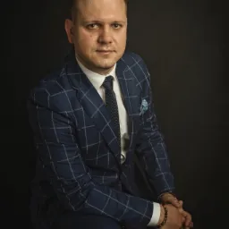 Portret mężczyzny w niebieskim garniturze w kratę, z poszetką w kieszonce, siedzącego na tle ciemnego tła, z bransoletką z kamieni na nadgarstku.