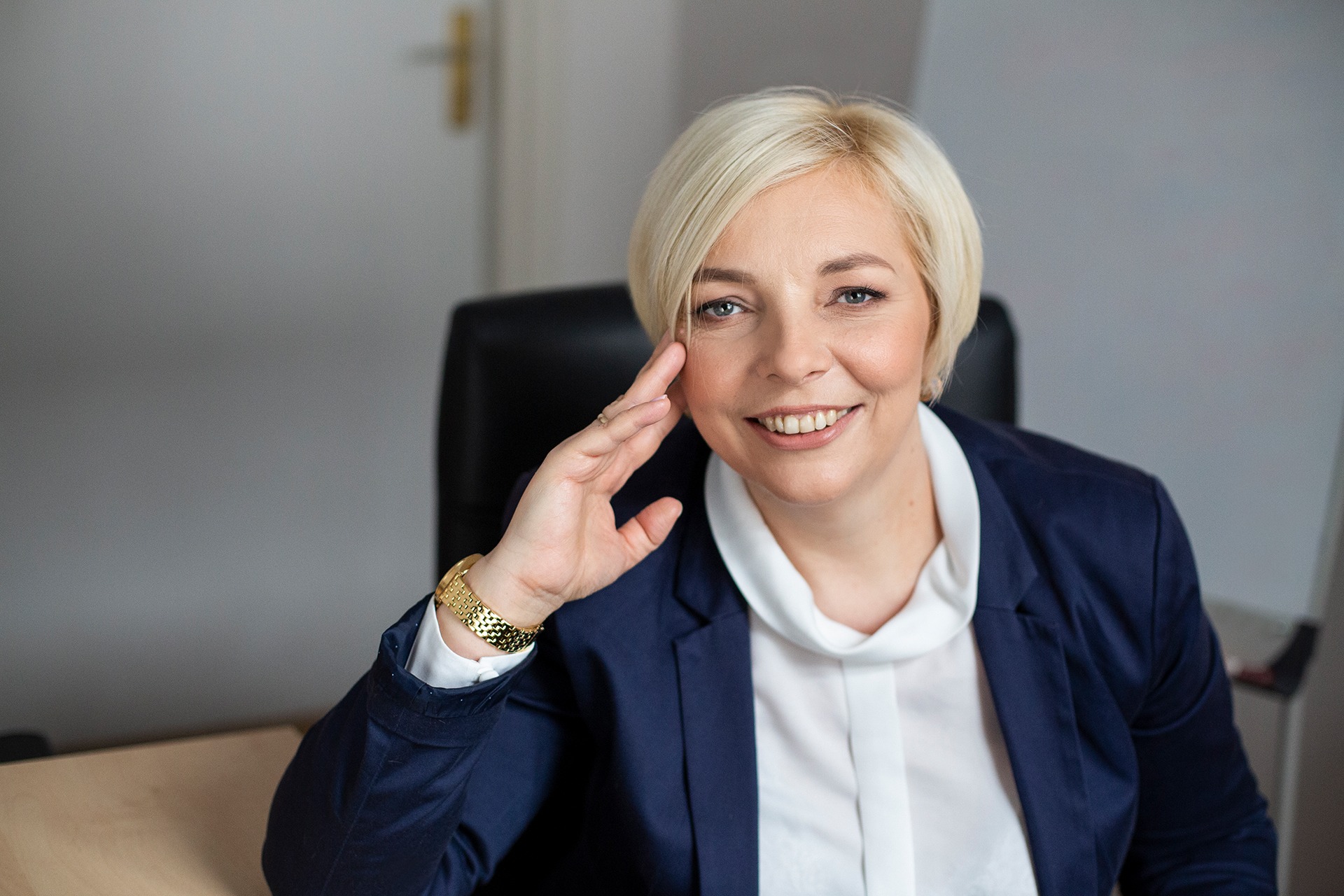 Portret uśmiechniętej blondynki w niebieskiej marynarce, z złotym zegarkiem na ręce, siedzącej przy biurku, sugerujący profesjonalizm i otwartość.