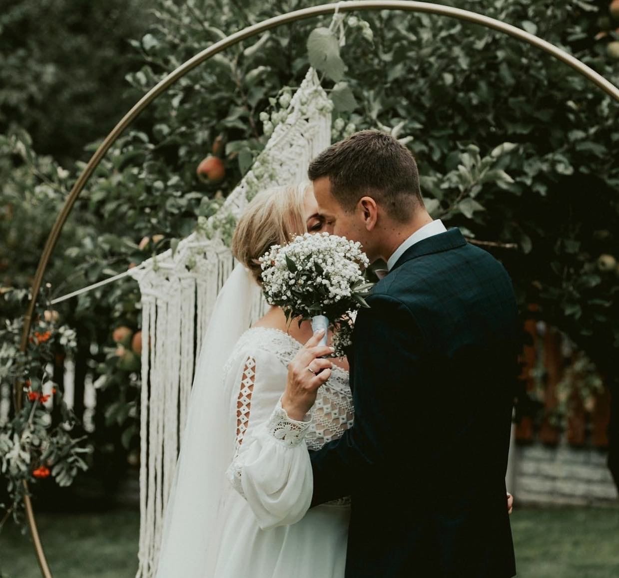 Para młoda całuje się za bukietem białych kwiatów przed okrągłym łukiem dekorowanym makramą i zielenią w ogrodzie.
