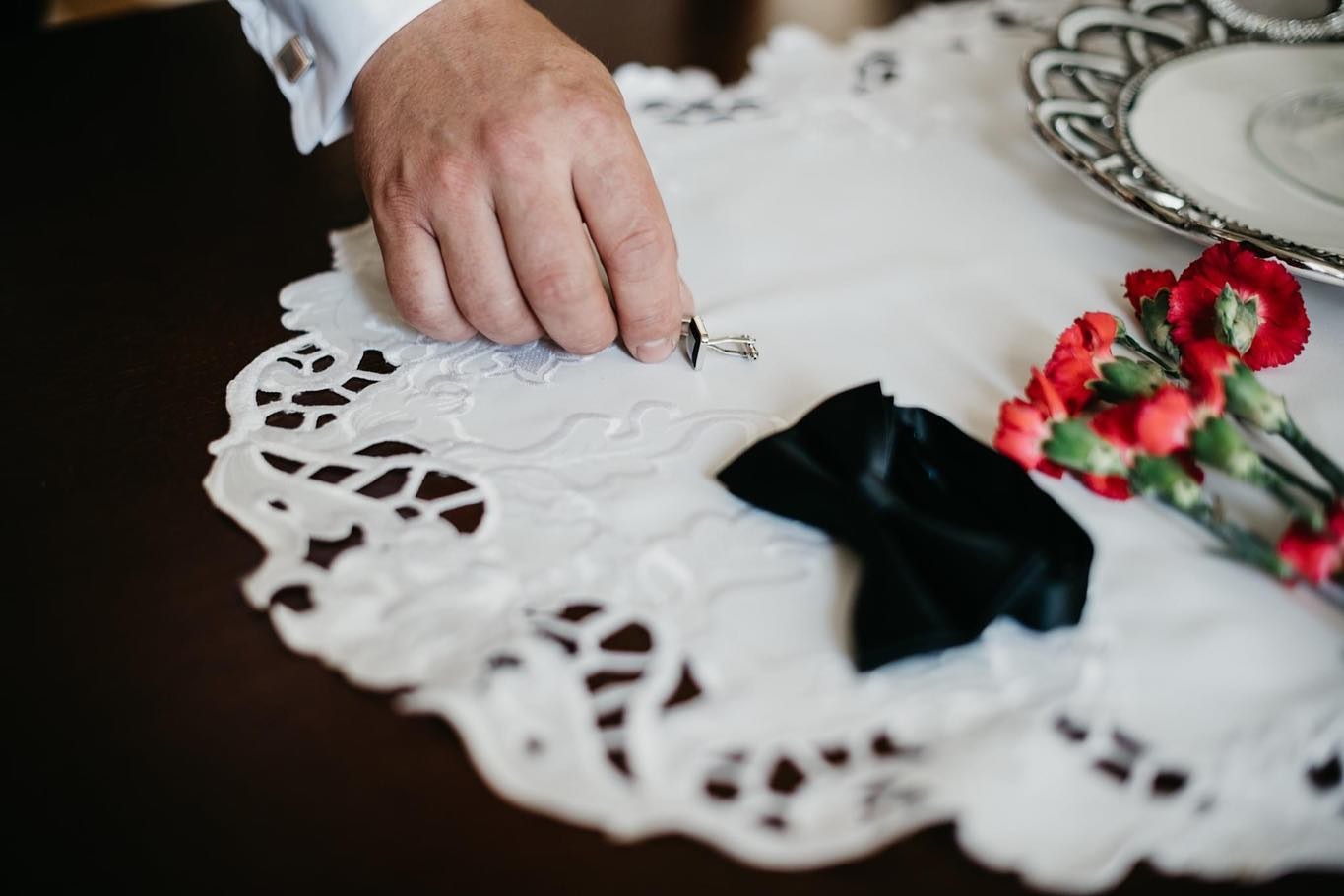 Przygotowania do ślubu: męska dłoń układa spinki do mankietów obok czarnej muchy i czerwonych goździków na białej, ażurowej serwetce.