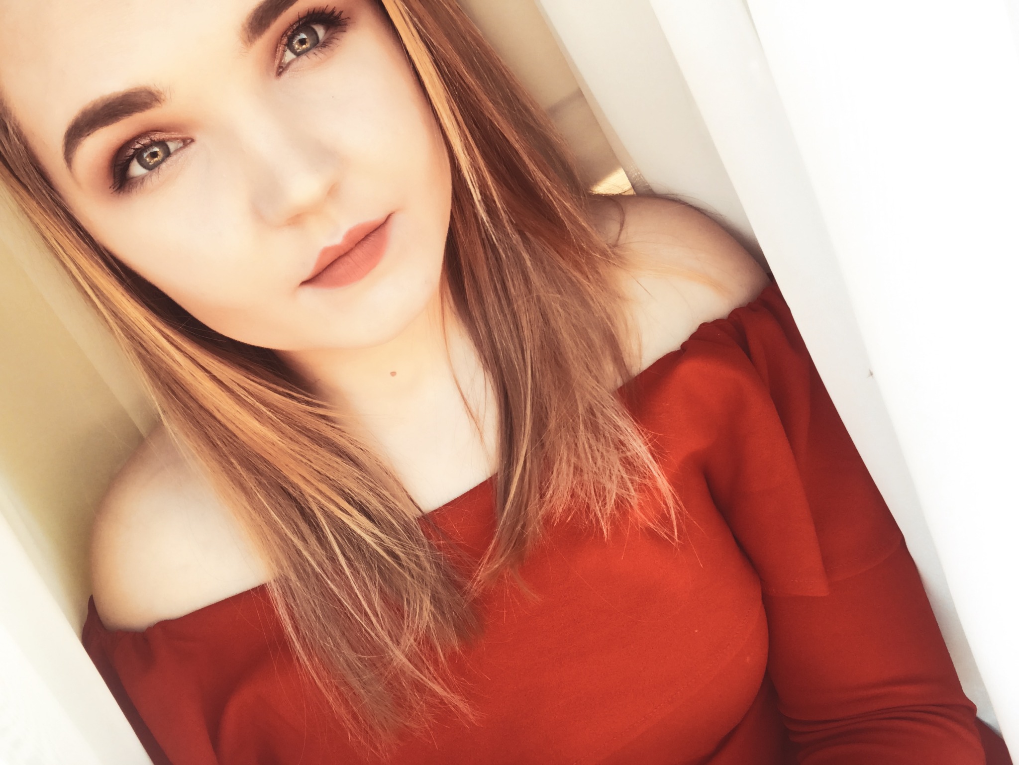 Portret kobiety z długimi blond włosami, w czerwonej bluzce z odkrytymi ramionami, patrzącej w górę.