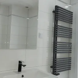 Nowoczesna łazienka z grzejnikiem drabinkowym w kolorze antracytowym, umywalką na szafce w odcieniach bieli i lustrem z oświetleniem LED.