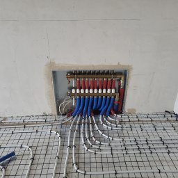 HYDRO-BUD Instalacje - Rozdzielacz ogrzewania podłogowego z niebieskimi i czerwonymi rurami w Podbrzeziu Dolnym, widok na siatkę montażową i surową ścianę.