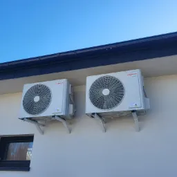 Dwa identyczne, białe klimatyzatory Viessmann zamontowane na wspornikach na elewacji budynku z czarnym oknem i rynną na tle błękitnego nieba.