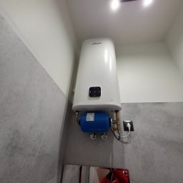 HYDRO-BUD Instalacje - Biały, wiszący bojler elektryczny Thermoval z niebieskim osprzętem, zamontowany na szarej ścianie w łazience. Widoczne przyłącza i gniazdko elektryczne.