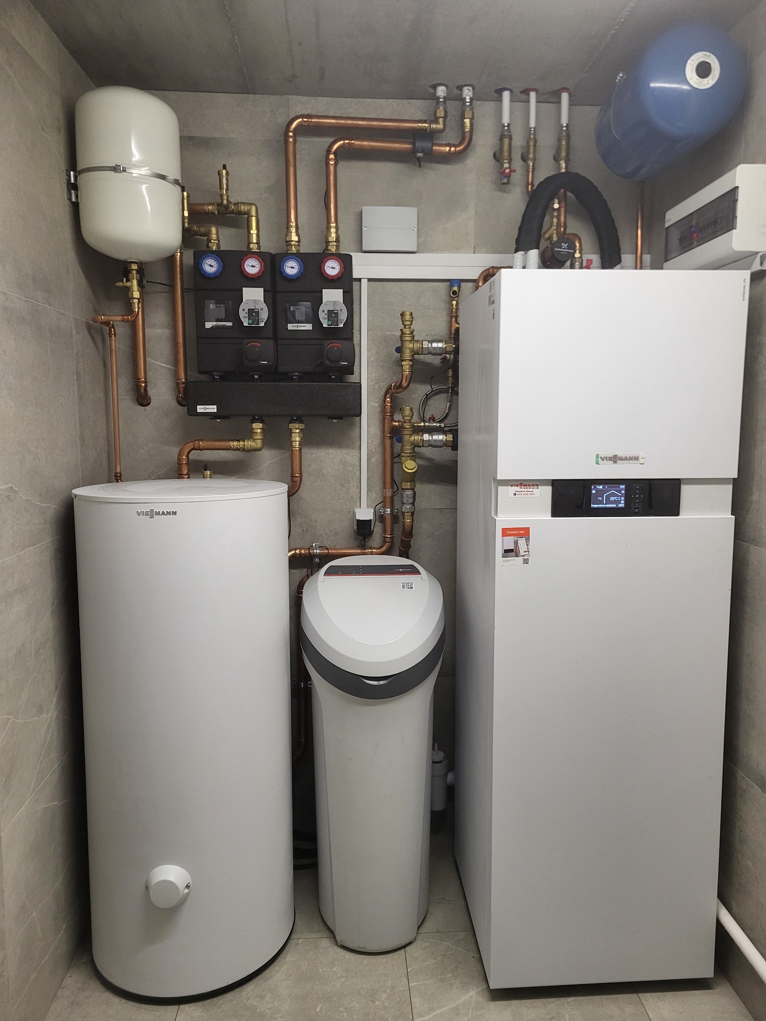 Kotłownia z nowoczesnym piecem grzewczym Viessmann, zasobnikiem ciepłej wody i zmiękczaczem wody, połączone miedzianą instalacją, na tle szarych płytek.