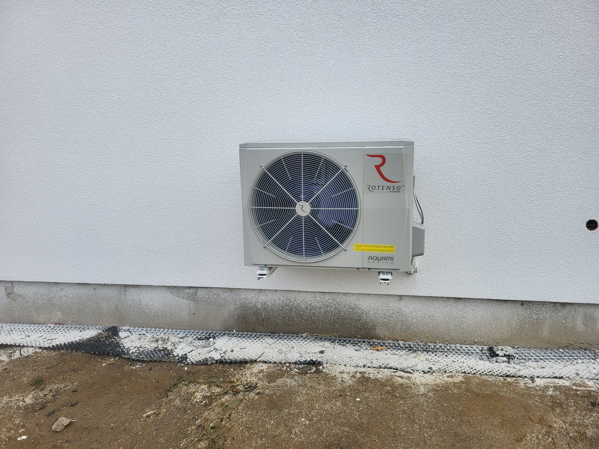 Szara zewnętrzna jednostka pompy ciepła Rotenso Aquami Series zamontowana na białej elewacji budynku, widoczne mocowania i fragment gruntu.