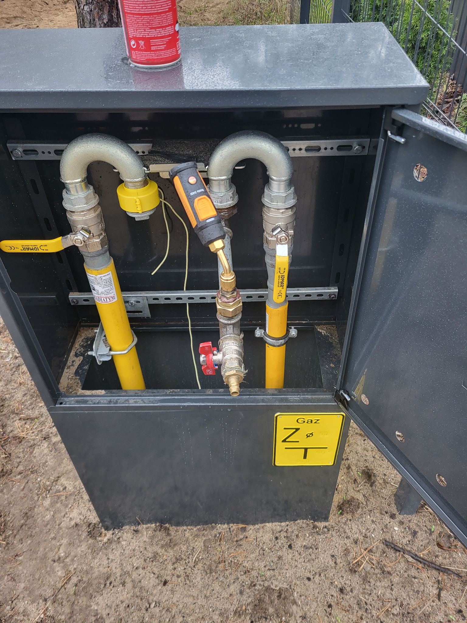 Szara szafka gazowa z otwartymi drzwiczkami, widoczne żółte rury gazowe z zaworami, przyłącza, miernik i puszka z pianką na górze.
