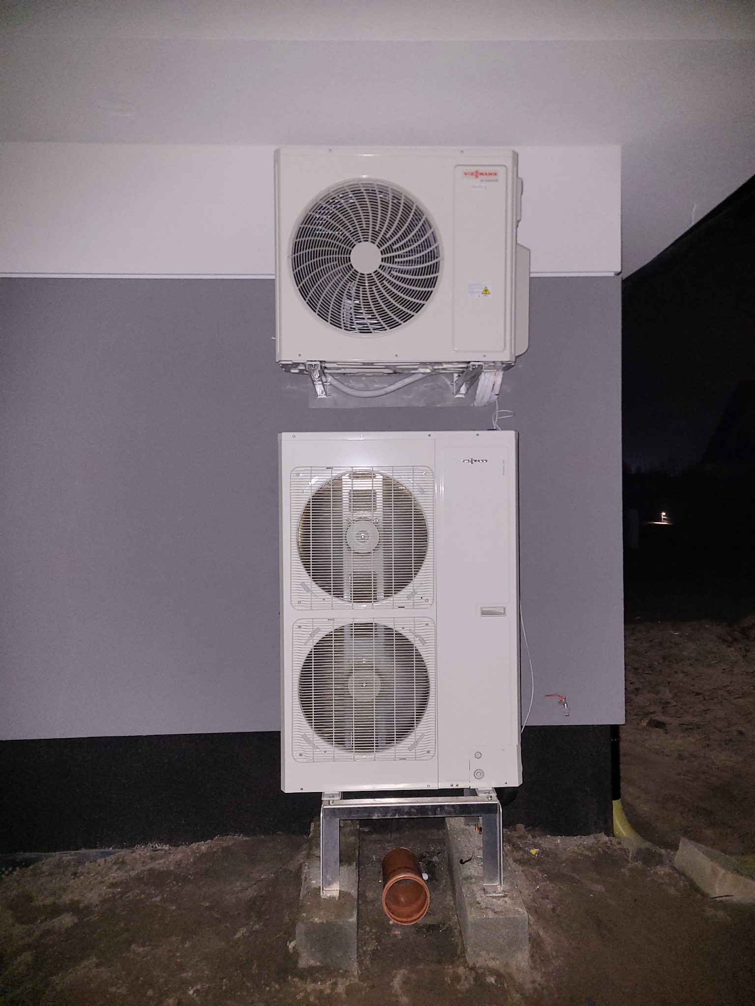 Dwa zewnętrzne moduły pompy ciepła zamontowane na szarej ścianie budynku, widoczny fragment rury odprowadzającej skropliny.