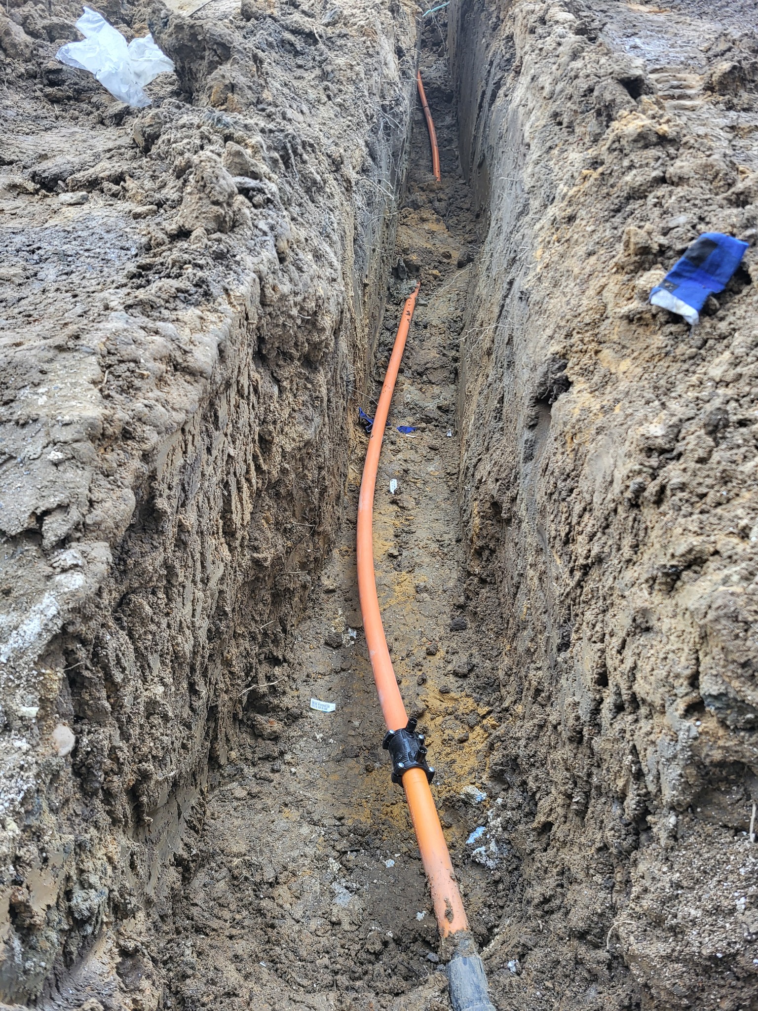 Wykop w ziemi z pomarańczową rurą instalacyjną z czarnymi złączkami, widoczna niebieska rękawica robocza na brzegu wykopu.
