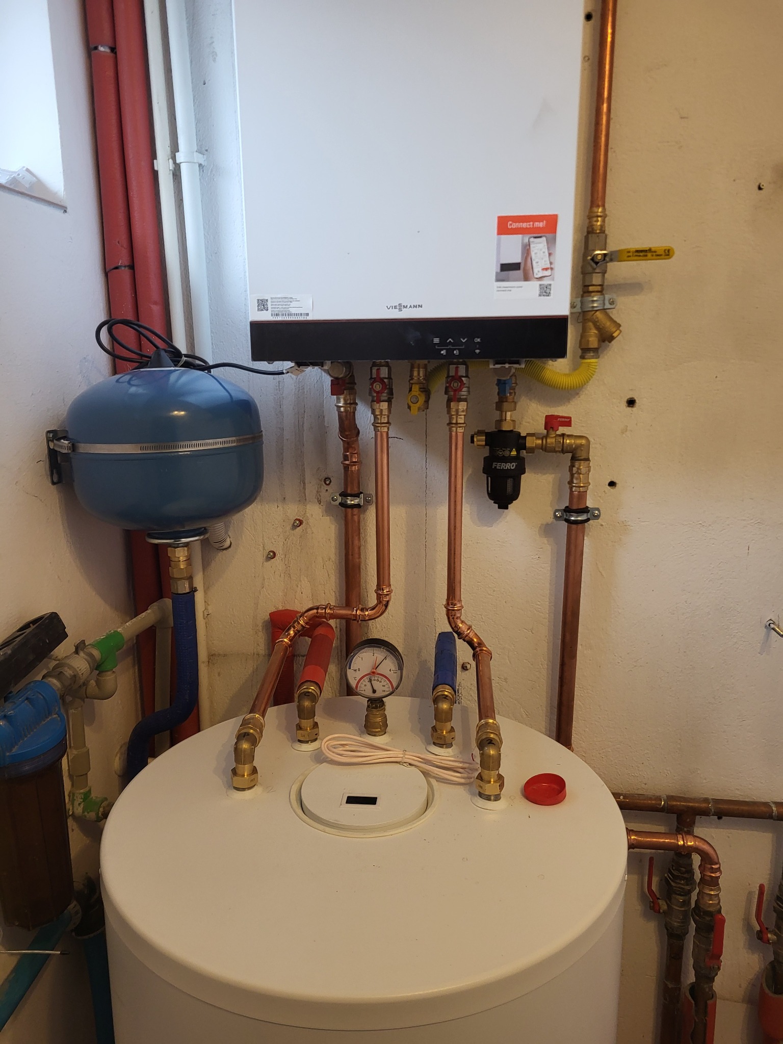 Nowoczesny piec gazowy Viessmann z podłączeniami z miedzi, zaworem Ferro i niebieskim zbiornikiem wyrównawczym, zamontowany na ścianie w pomieszczeniu technicznym.