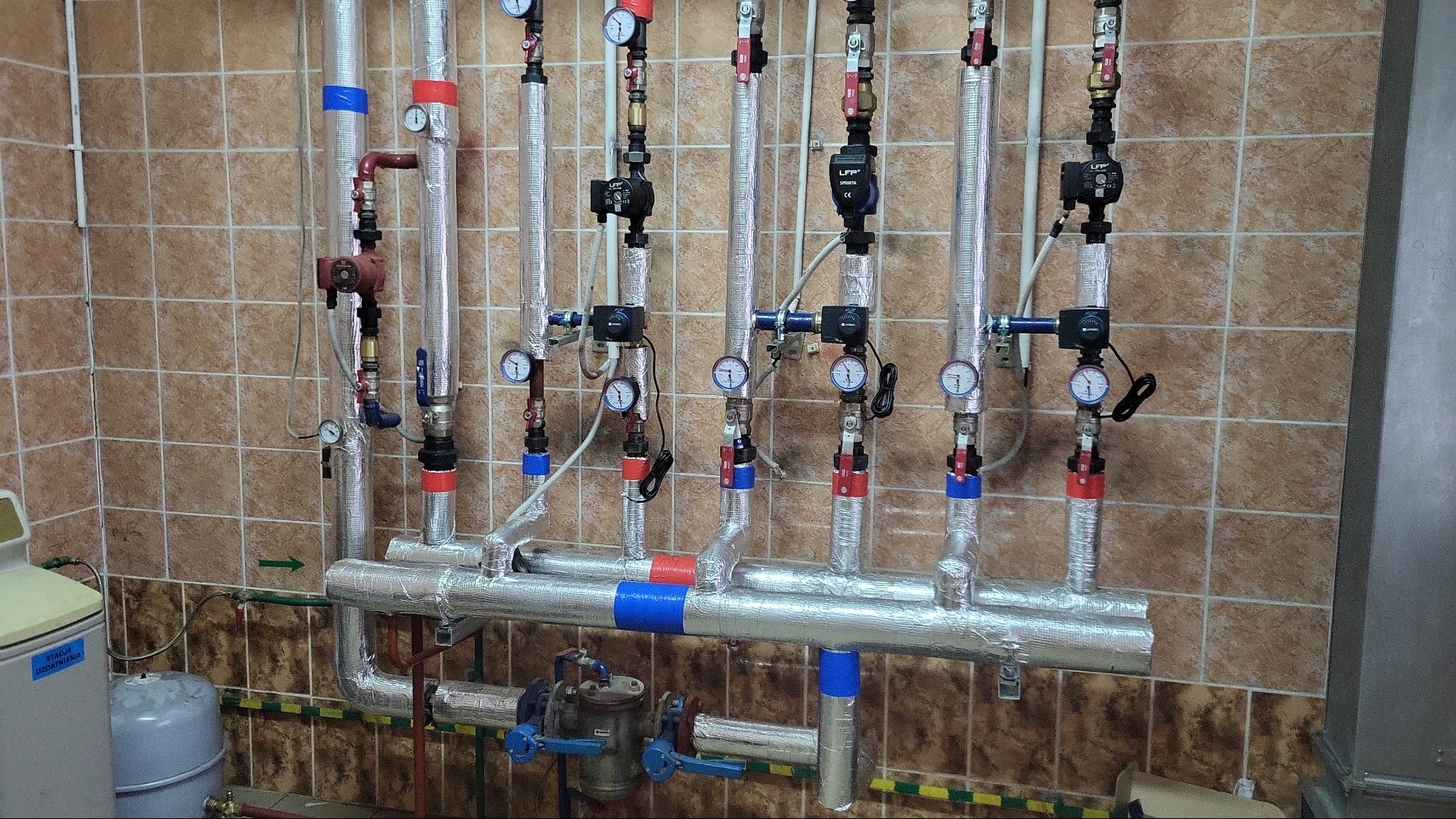 Rozdzielacz instalacji grzewczej z kilkoma obiegami, każdy z pompą obiegową i manometrami, izolowany folią aluminiową, na tle brązowych płytek.