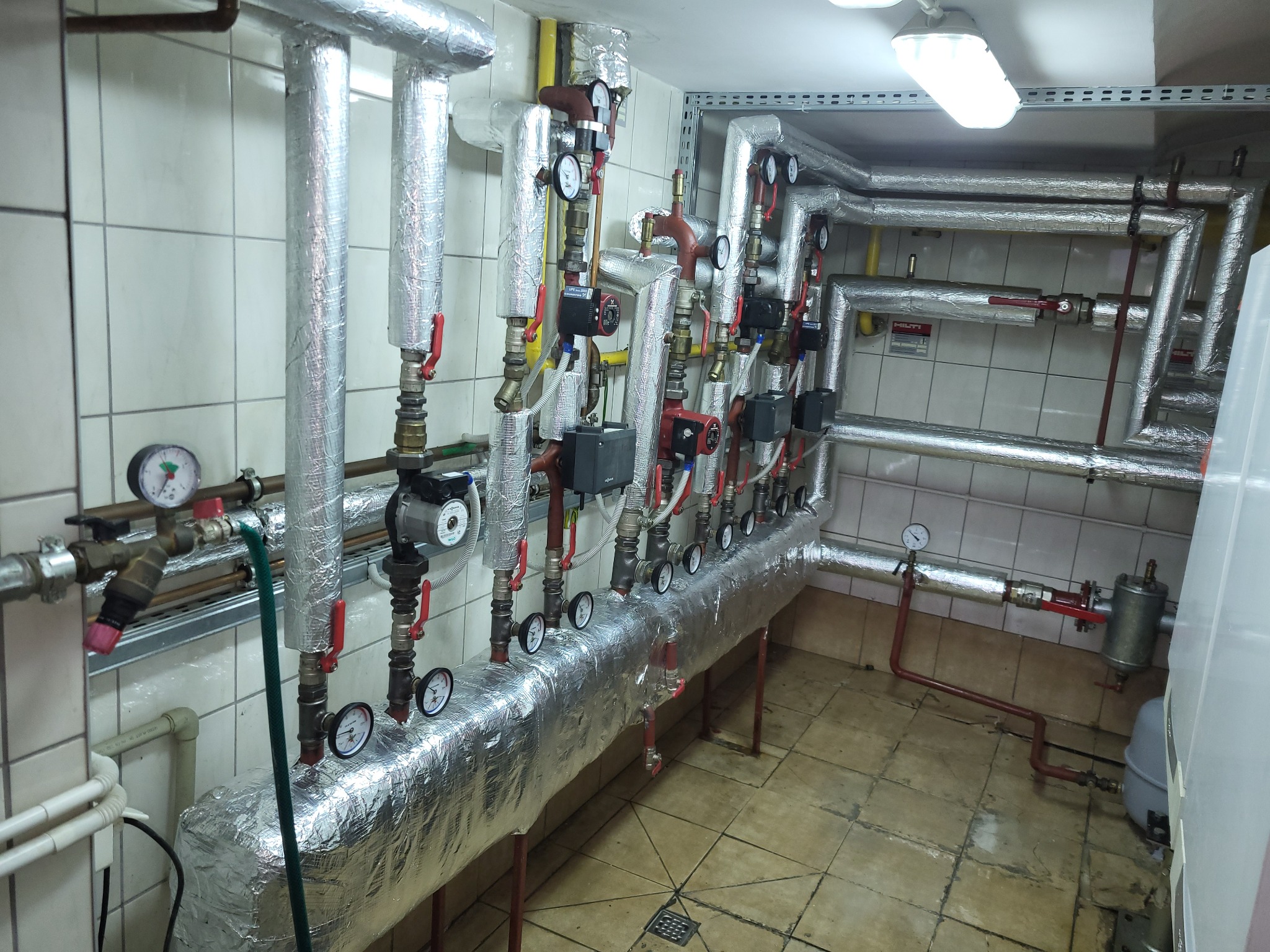 Węzeł cieplny z izolowanymi rurami, manometrami i zaworami w pomieszczeniu technicznym z białymi kafelkami.