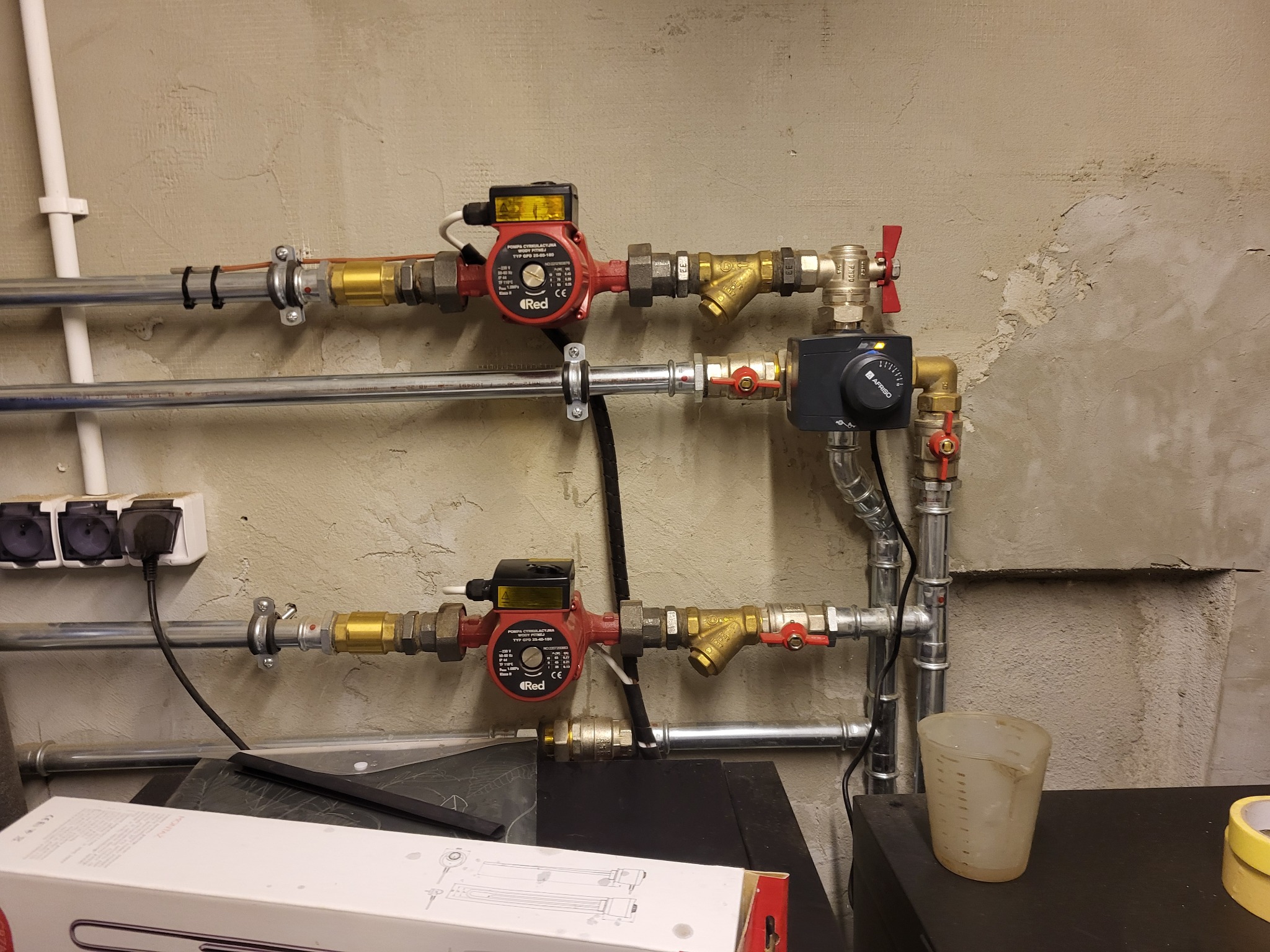 Dwie czerwone pompy obiegowe zamontowane na metalowych rurach z zaworami, widoczne elementy instalacji grzewczej, w tle surowa ściana, na pierwszym planie plastikowy kubek z miarką.